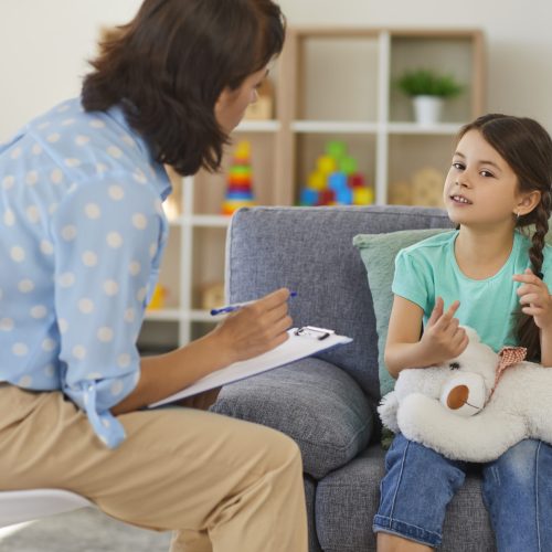 Supportive psychologist with clipboard listening to little child during therapy session. Preschool girl feeling at ease in therapist's office sharing her thoughts and concerns