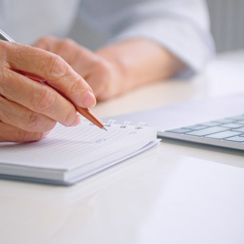 Old woman wrinkly hands write in paper notebook with pen at table near grey laptop keyboard Old woman wrinkly hands write in paper notebook with pen at table near grey laptop keyboard